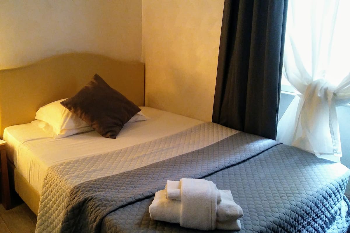 Una cama bien hecha con una almohada, una mesita de noche con una lámpara y una ventana con cortinas.