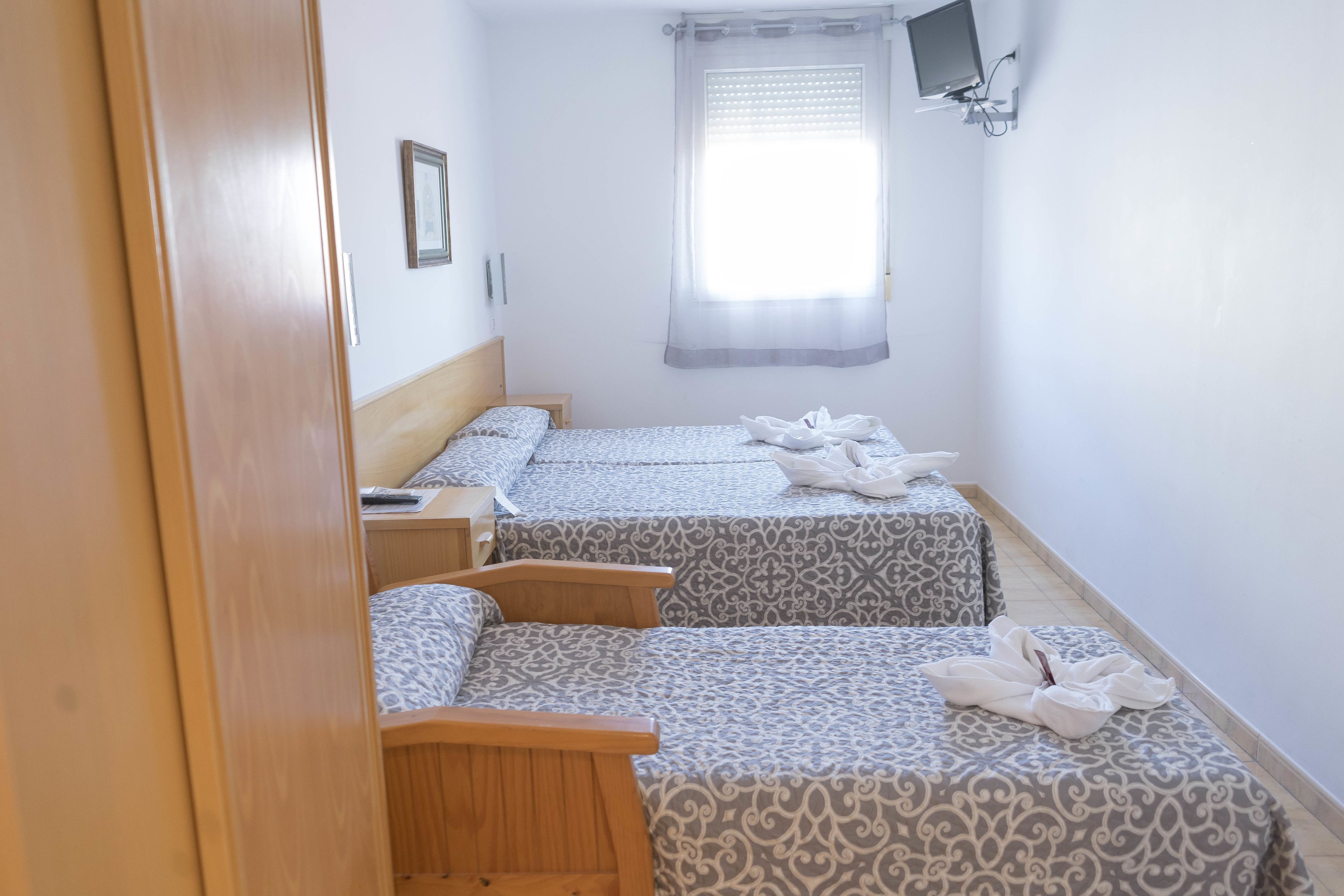 Une chambre avec deux lits, une porte en bois, une petite table de chevet, un téléviseur fixé au mur et une fenêtre avec des rideaux.