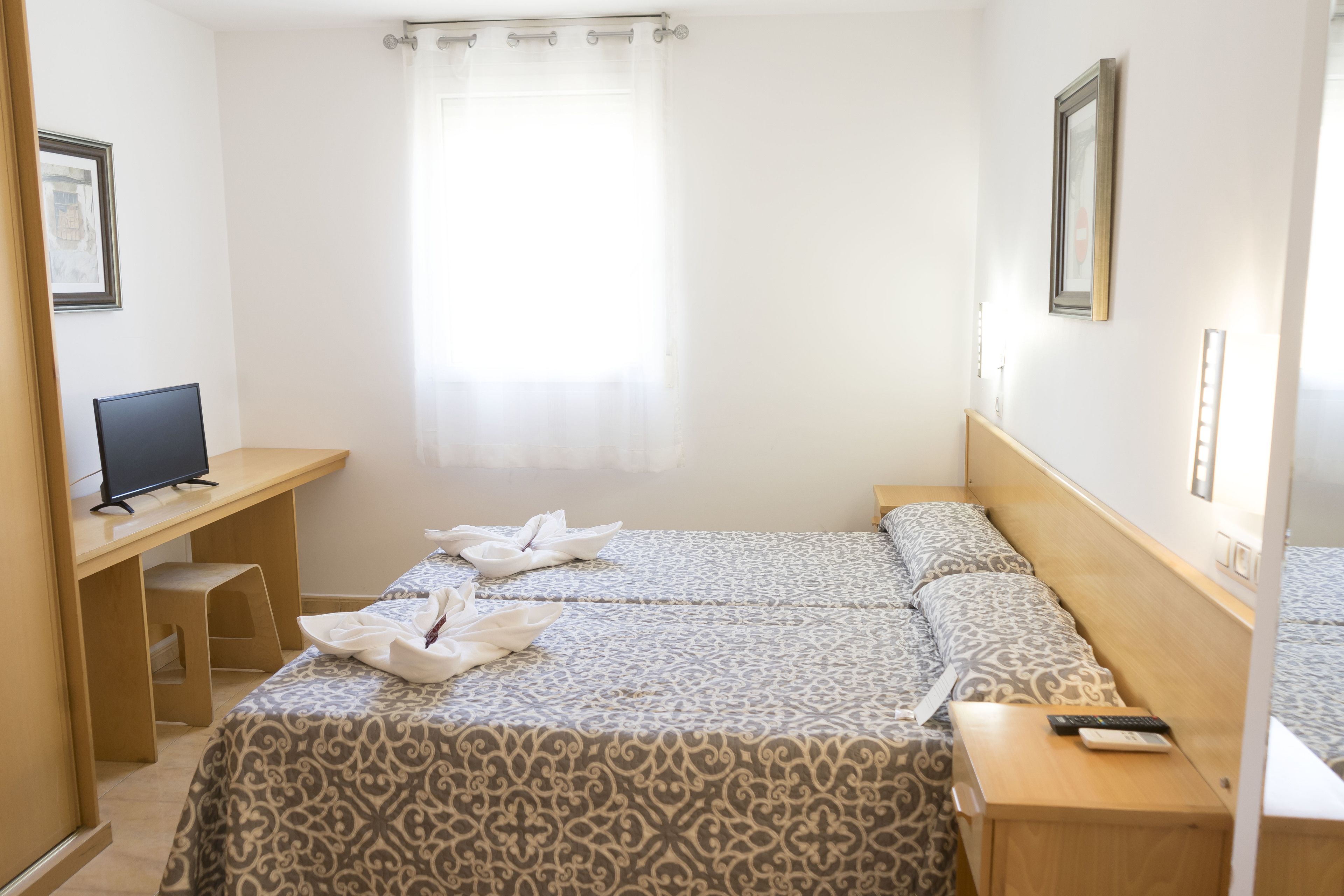 A hotel room with two beds, a desk, a chair, a TV, and a window with curtains.
