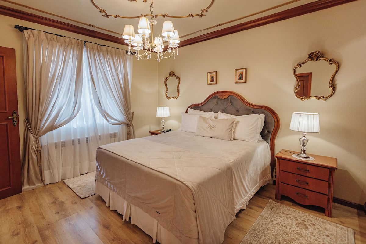A bedroom with a large bed, a wooden nightstand, a chandelier, and a mirror on the wall.