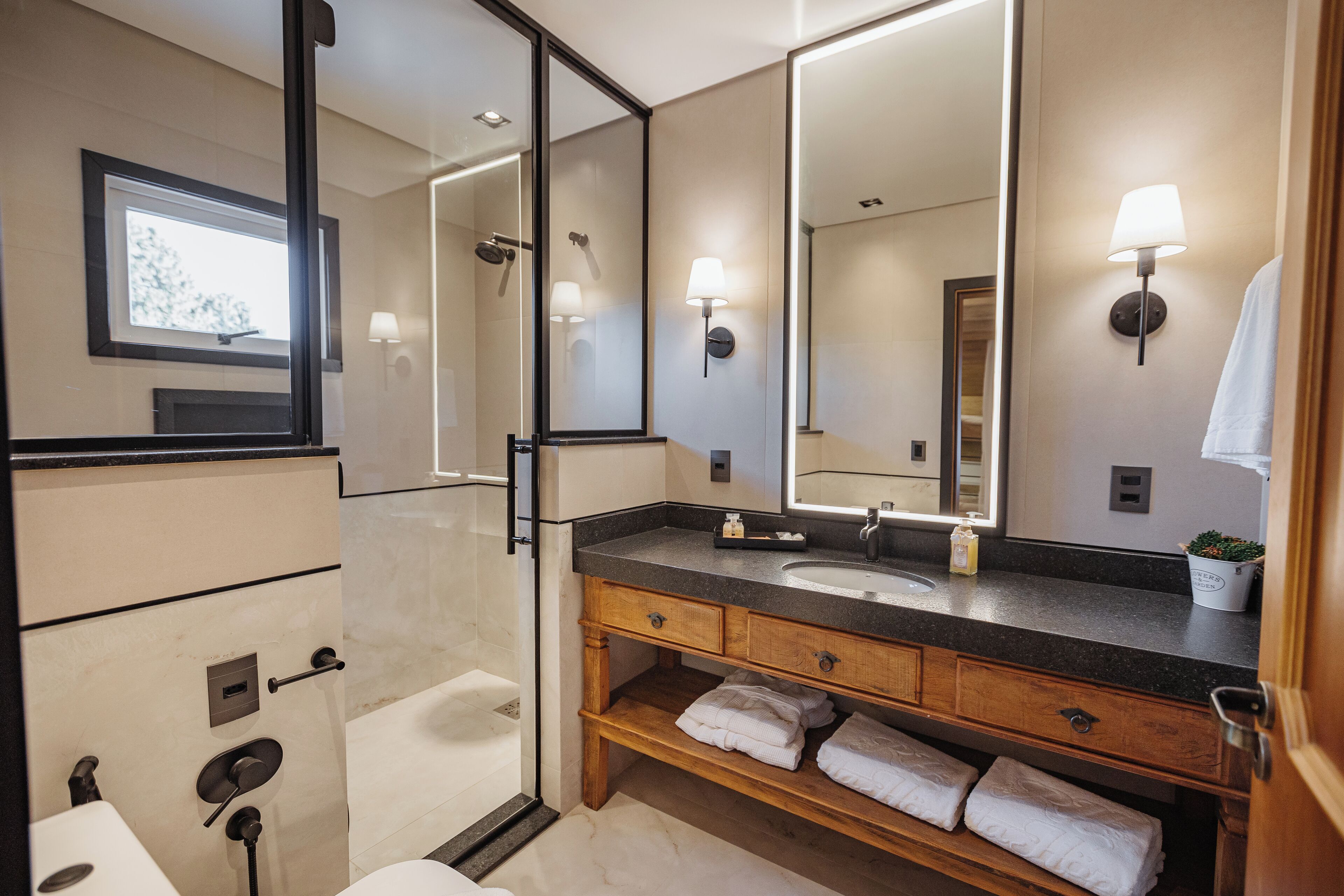 Un baño moderno con un espejo grande, un lavabo y una ducha. Hay toallas en un estante y una ventana con vista a árboles.