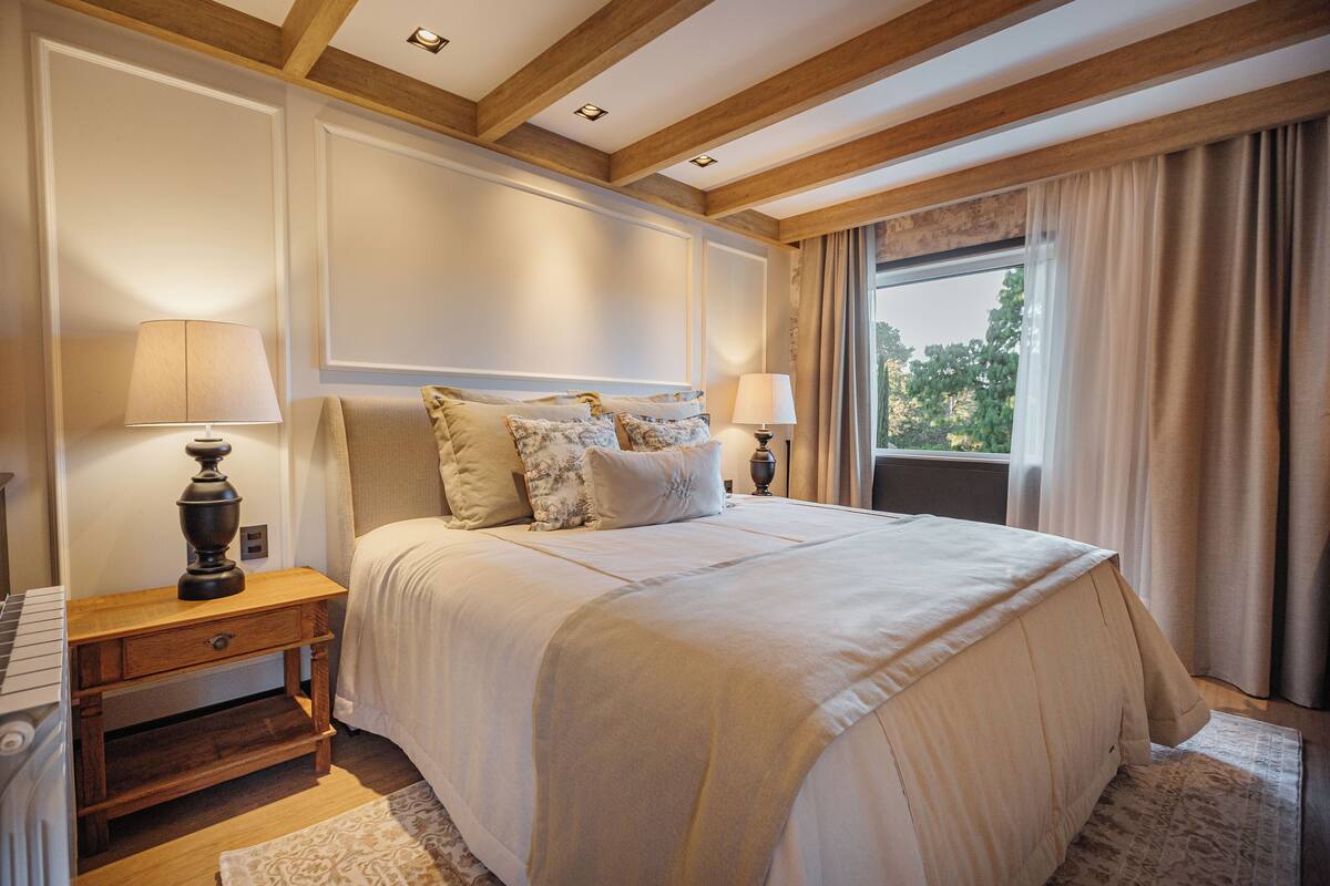 A large bed with a headboard, two bedside tables with lamps, a wooden nightstand, and a view of the outdoors through a window with curtains.