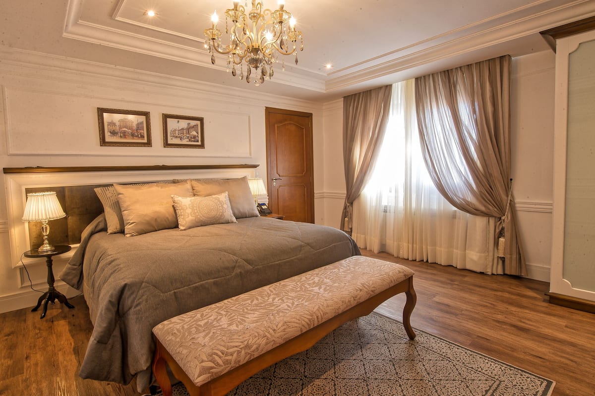 A bedroom with a large bed, a bench, a chandelier, and two framed pictures on the wall.