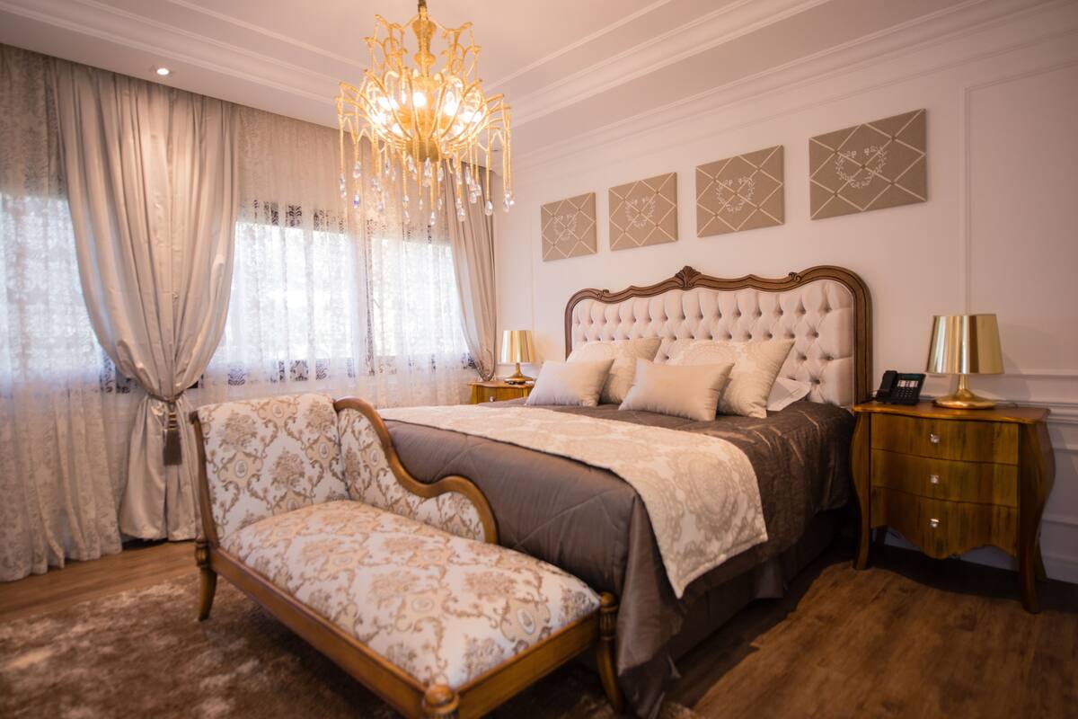 A bedroom with a large bed, a chandelier, a nightstand, and a chair.