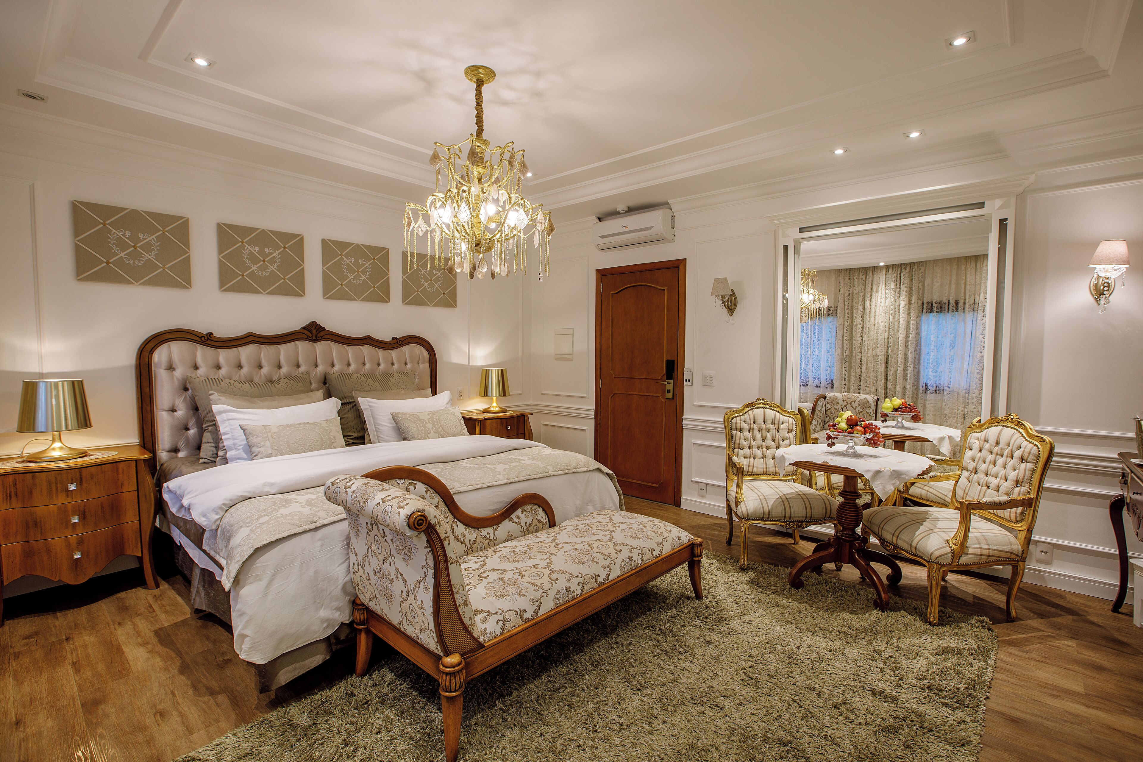 A bedroom with a large bed, a sitting area with chairs, a chandelier, and a dining table with chairs.