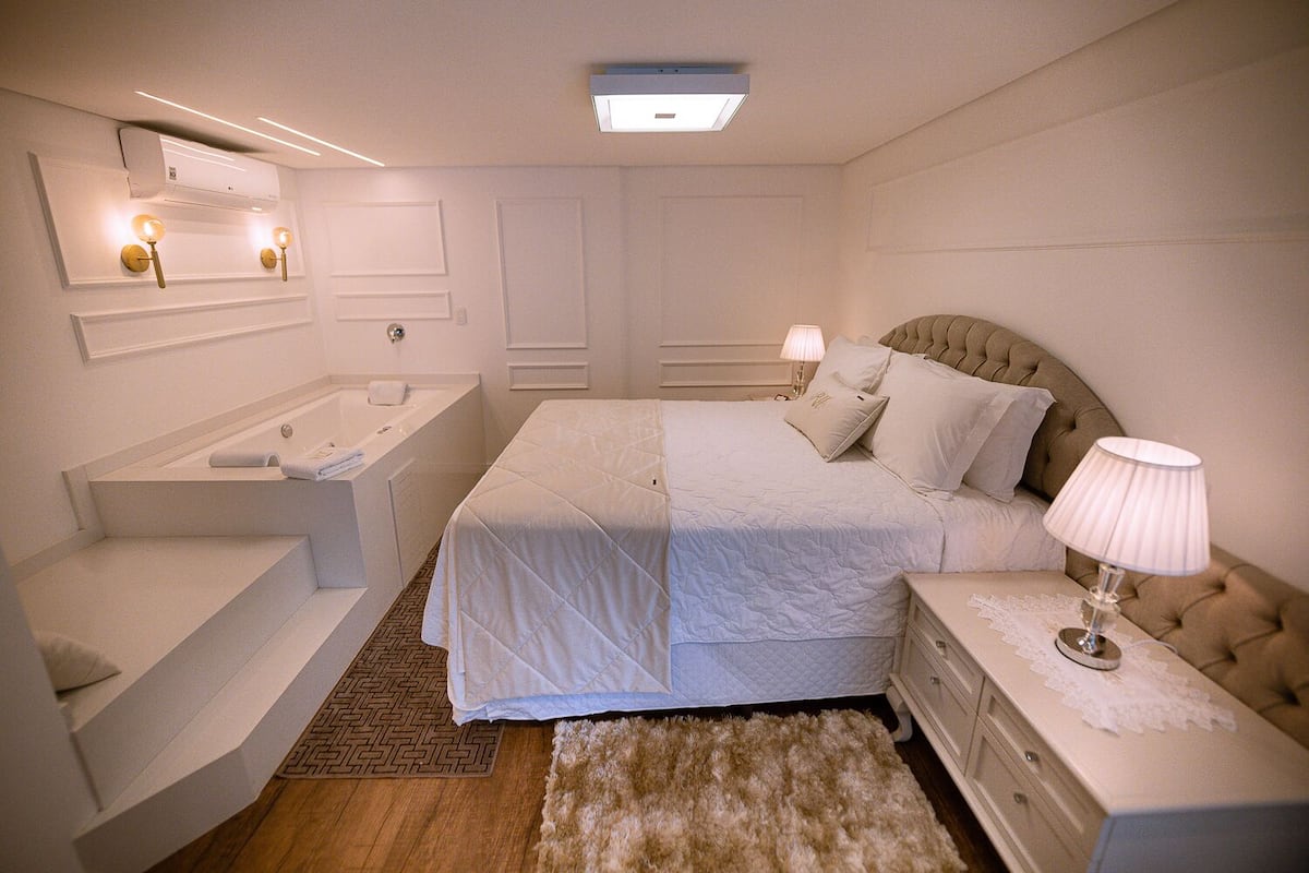 A modern bedroom with a large bed, a separate bathtub, a vanity with a mirror, and a bedside table with a lamp.