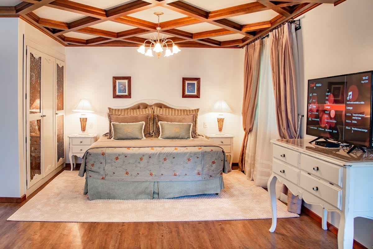 A bedroom with a wooden ceiling, a bed with floral bedding, two bedside lamps, a TV on the right, and a wooden wardrobe.