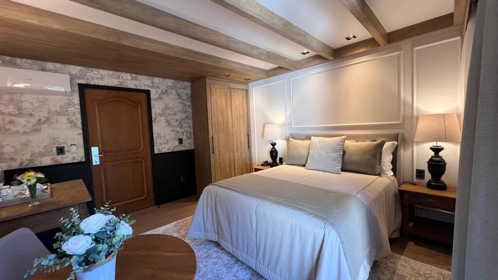 A bedroom with a large bed, wooden ceiling beams, a nightstand, and a desk with a lamp.