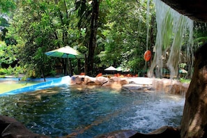 Un área tranquila de piscina al aire libre con cascada, rodeada de exuberante vegetación y protegida por sombrillas.