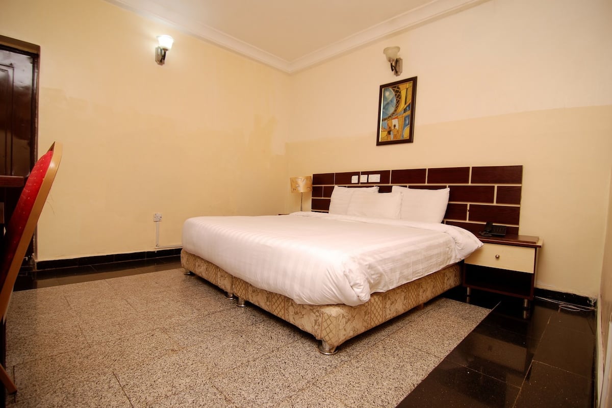 Quarto de hotel com uma cama grande, uma mesa de cabeceira, um quadro pendurado na parede e uma porta.