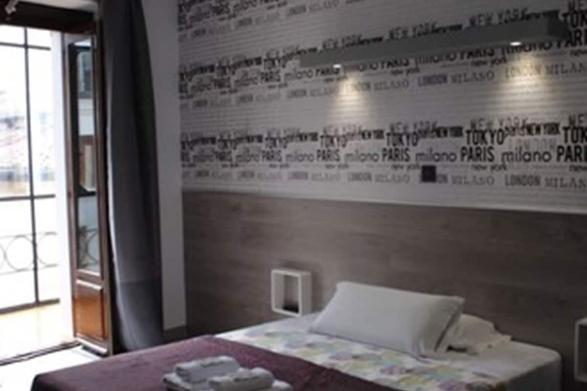 A hotel room with a bed, a wall with repeated city names, and a window with curtains.
