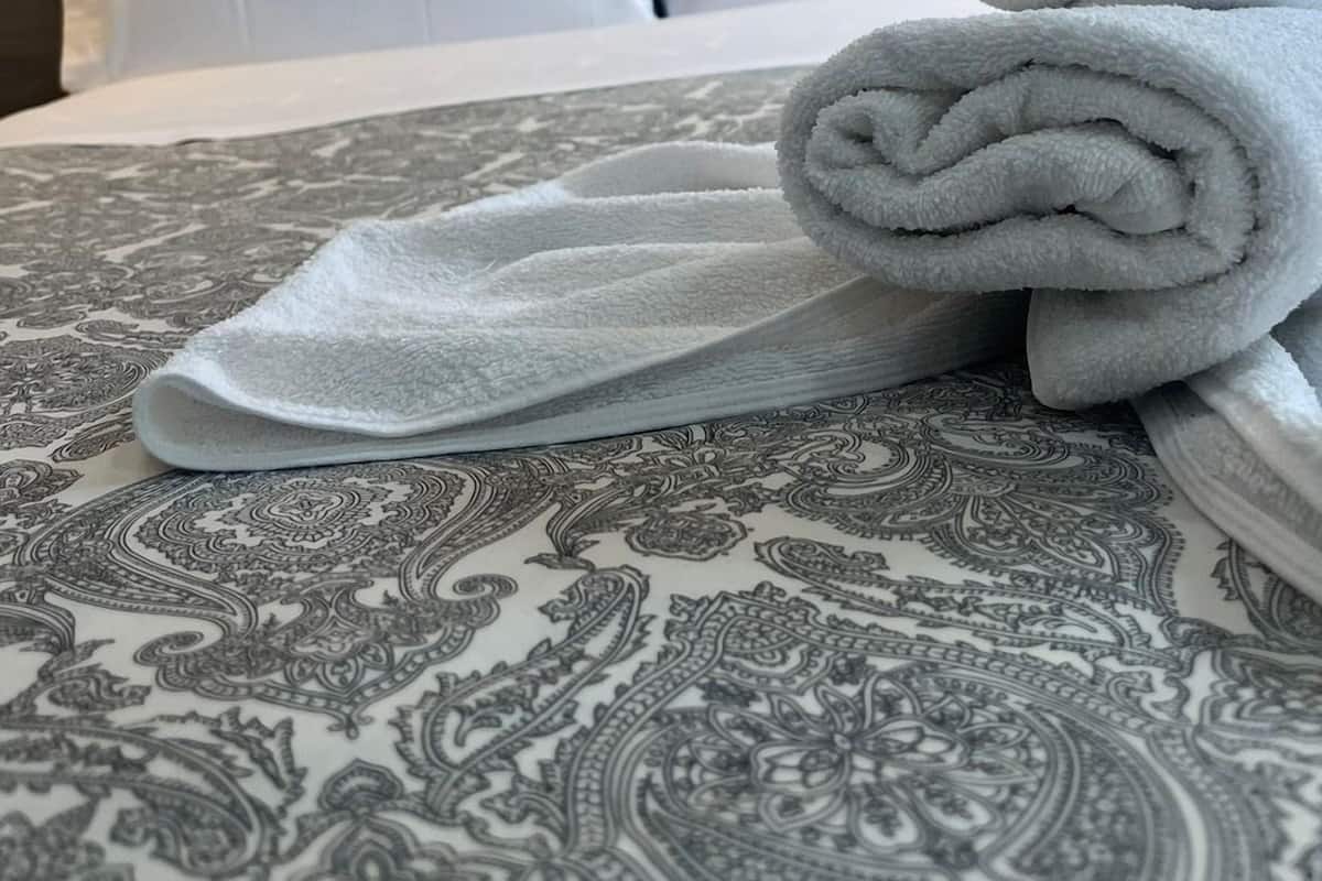 A hotel room with a bed, two white pillows, and a rolled-up towel on a patterned bedspread.