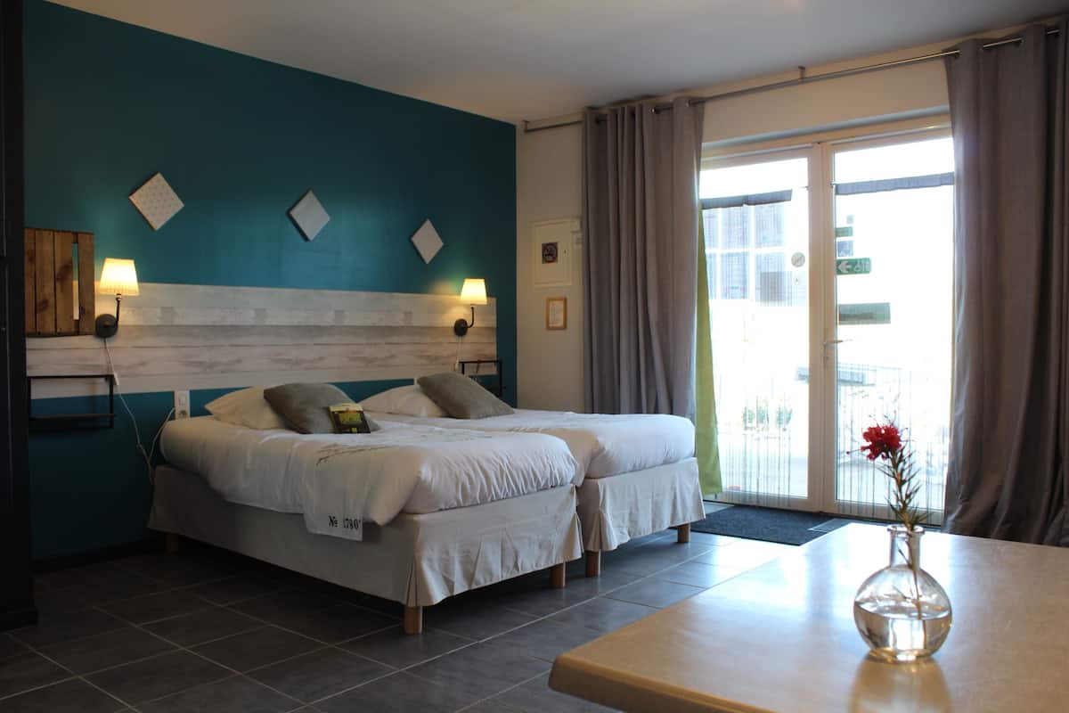 A hotel room with two beds, a wooden headboard, a small table with a vase, and a view of the outside through a glass door.
