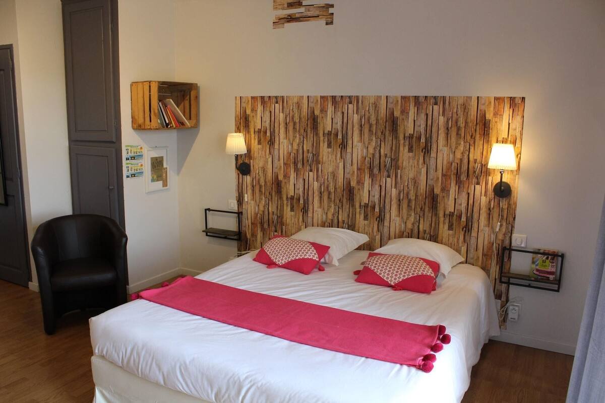 A bedroom with a wooden headboard, two bedside lamps, a bed with white bedding and red pillows, a black chair, and a wooden shelf with books.