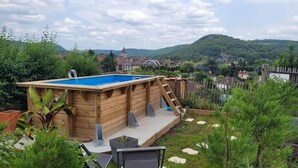 Una terraza de madera con piscina, rodeada de vegetación y vistas a un pueblo y colinas al fondo.