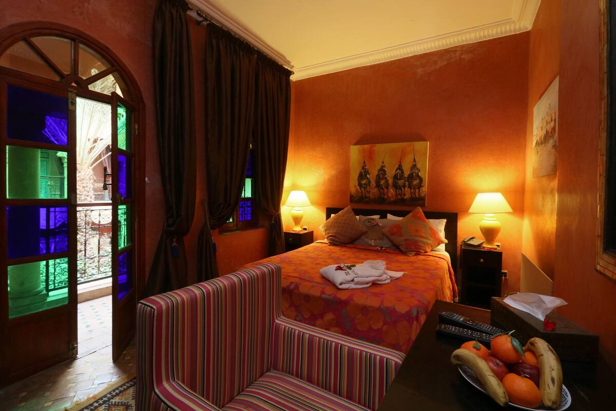 A hotel room with a bed, two bedside lamps, a nightstand, a chair, a fruit bowl, and a painting on the wall.