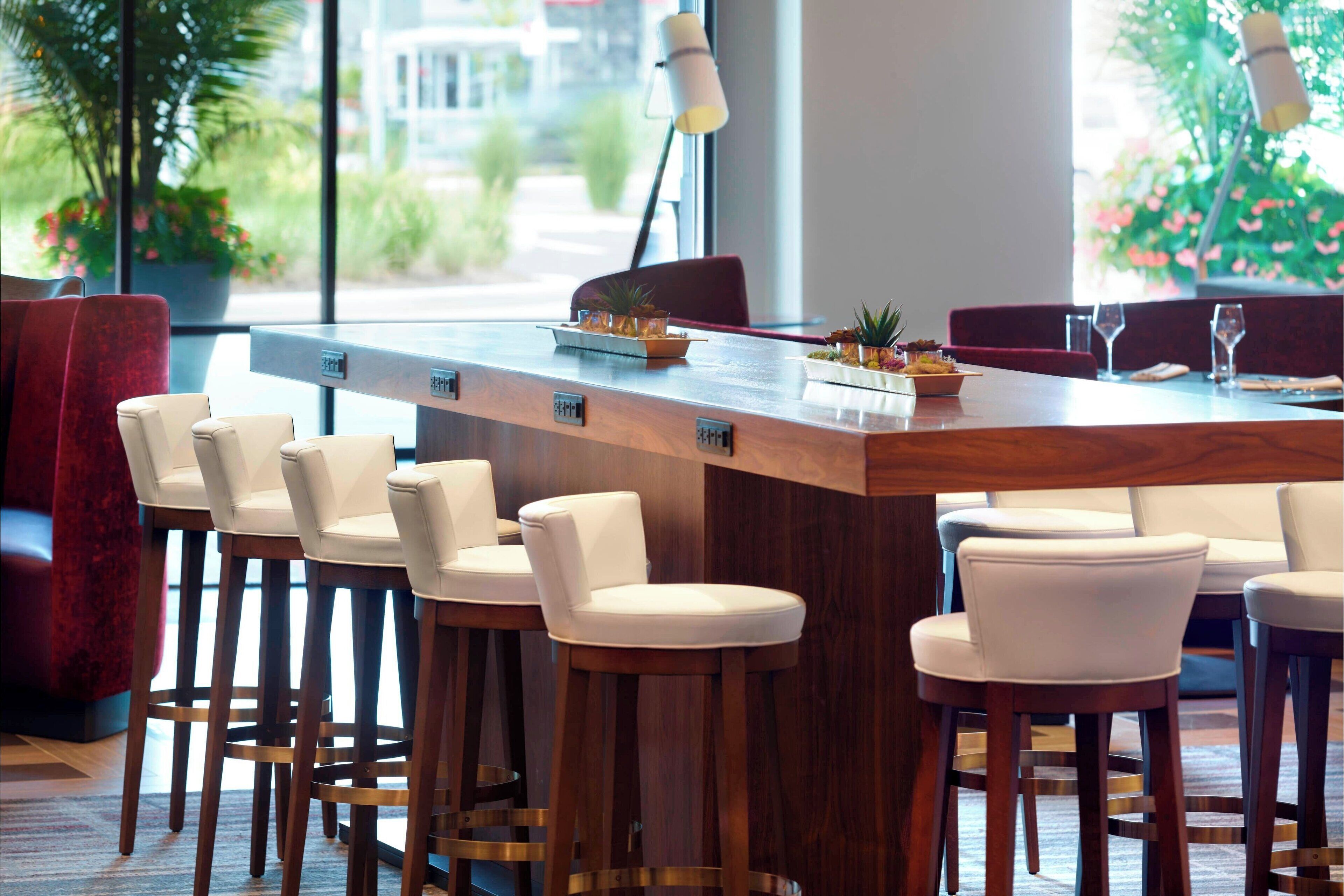 A modern bar area with wooden stools and a long counter, surrounded by red upholstered chairs.