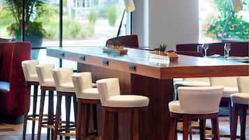 A modern bar area with wooden stools and a long counter, surrounded by red upholstered chairs.