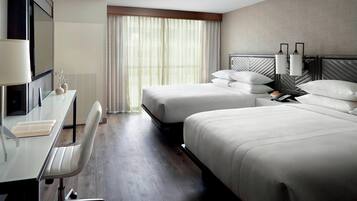 Ein Hotelzimmer mit zwei Betten, einem Schreibtisch mit Stuhl, einem Fernseher und einem Fenster mit Vorhängen.