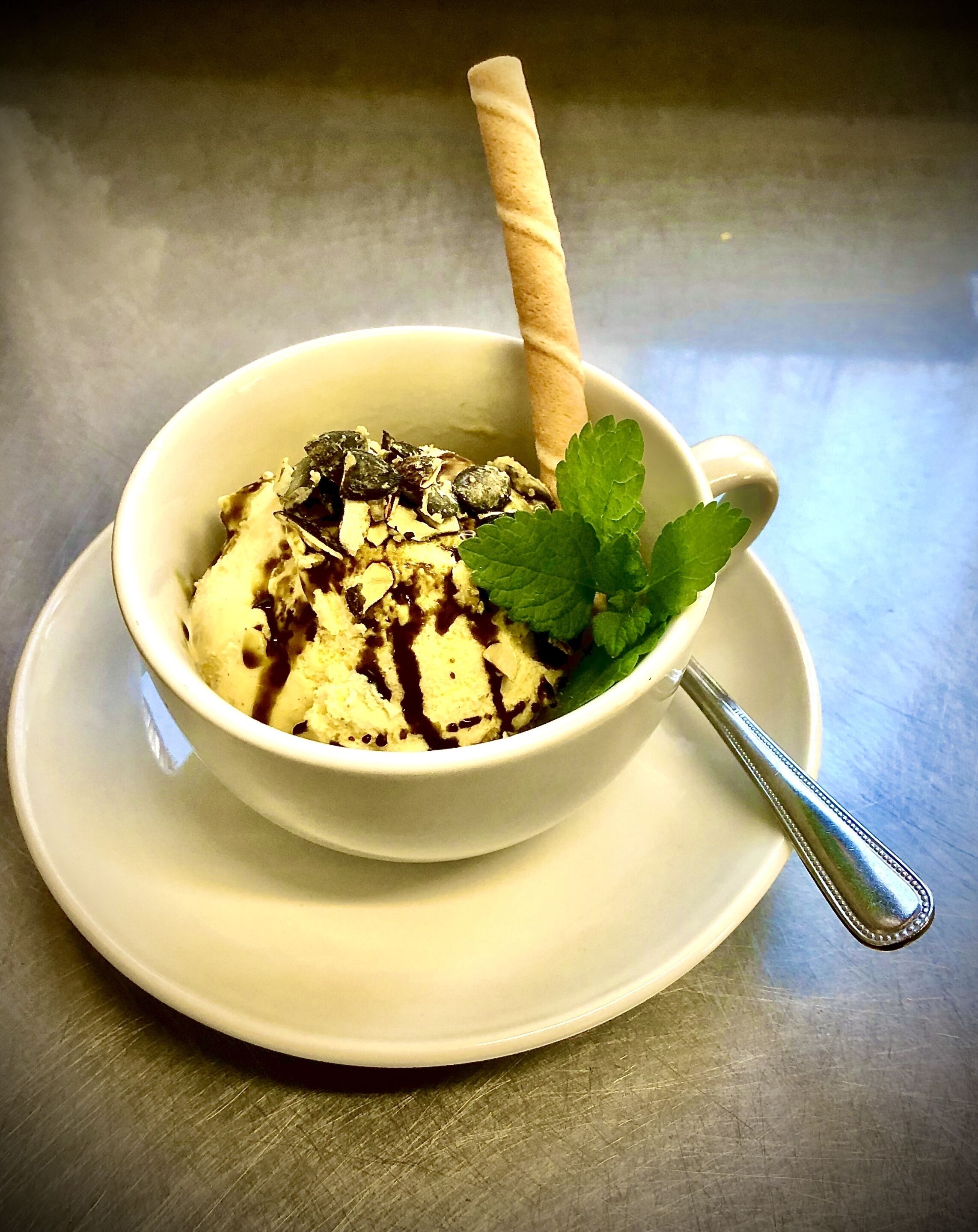 A bowl of ice cream with chocolate syrup and a mint leaf garnish on a white plate with a spoon.