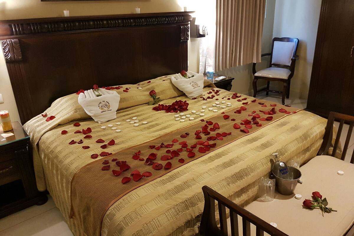 A neatly made bed with rose petals and candles, a wooden nightstand, a chair, and a table with a bucket and a bottle.