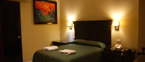 A hotel room with a bed, two bedside lamps, a nightstand, a telephone, and a painting on the wall.