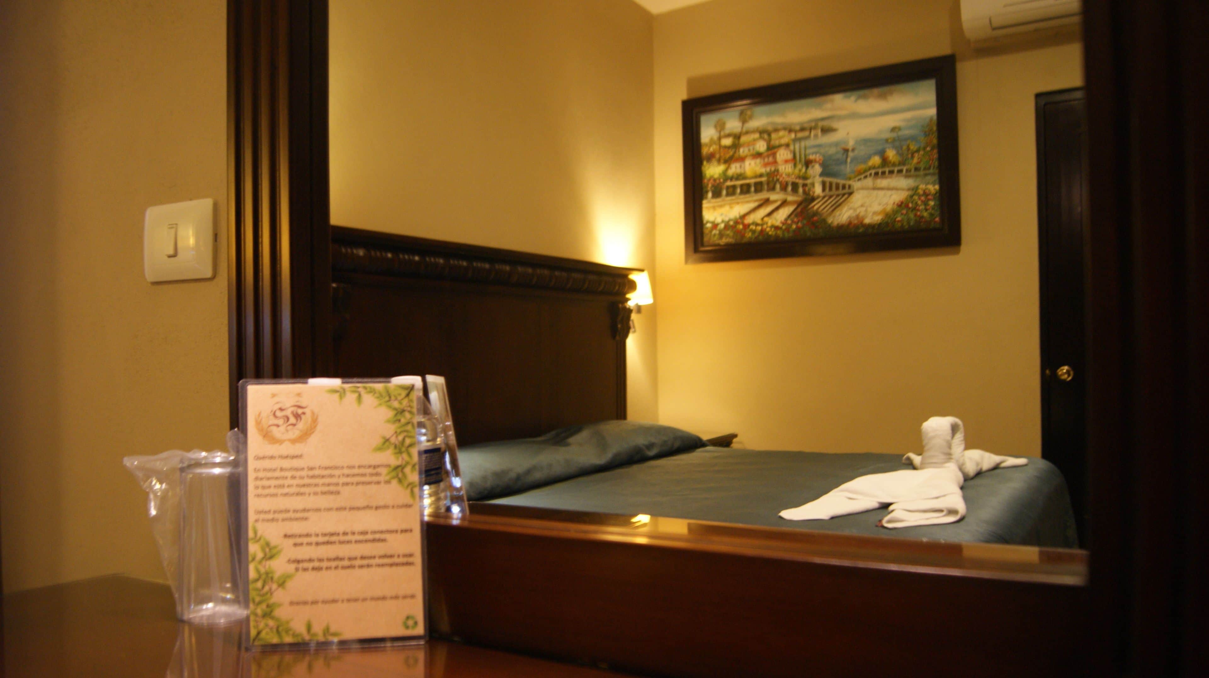 A hotel room with a wooden bed, a nightstand with a glass vase and a brochure, a painting on the wall, and a door.