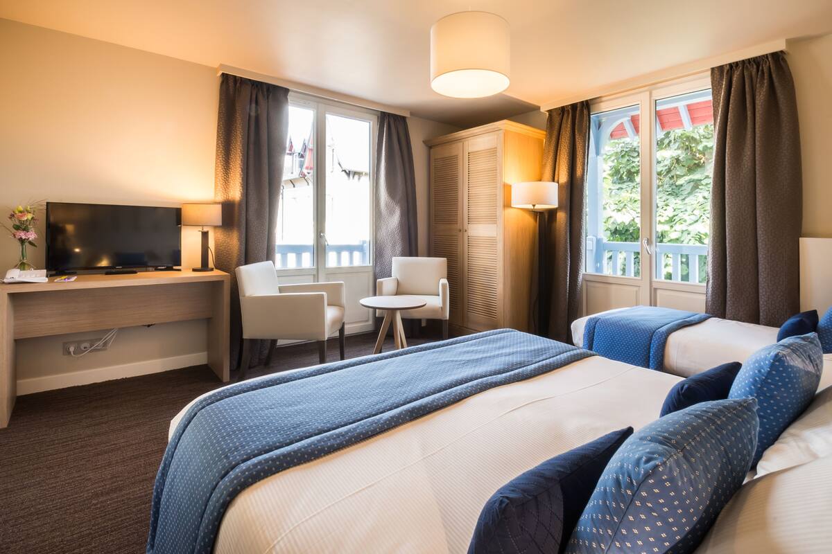 A hotel room with two beds, a desk with a TV, a chair, a small table, and a window with curtains.
