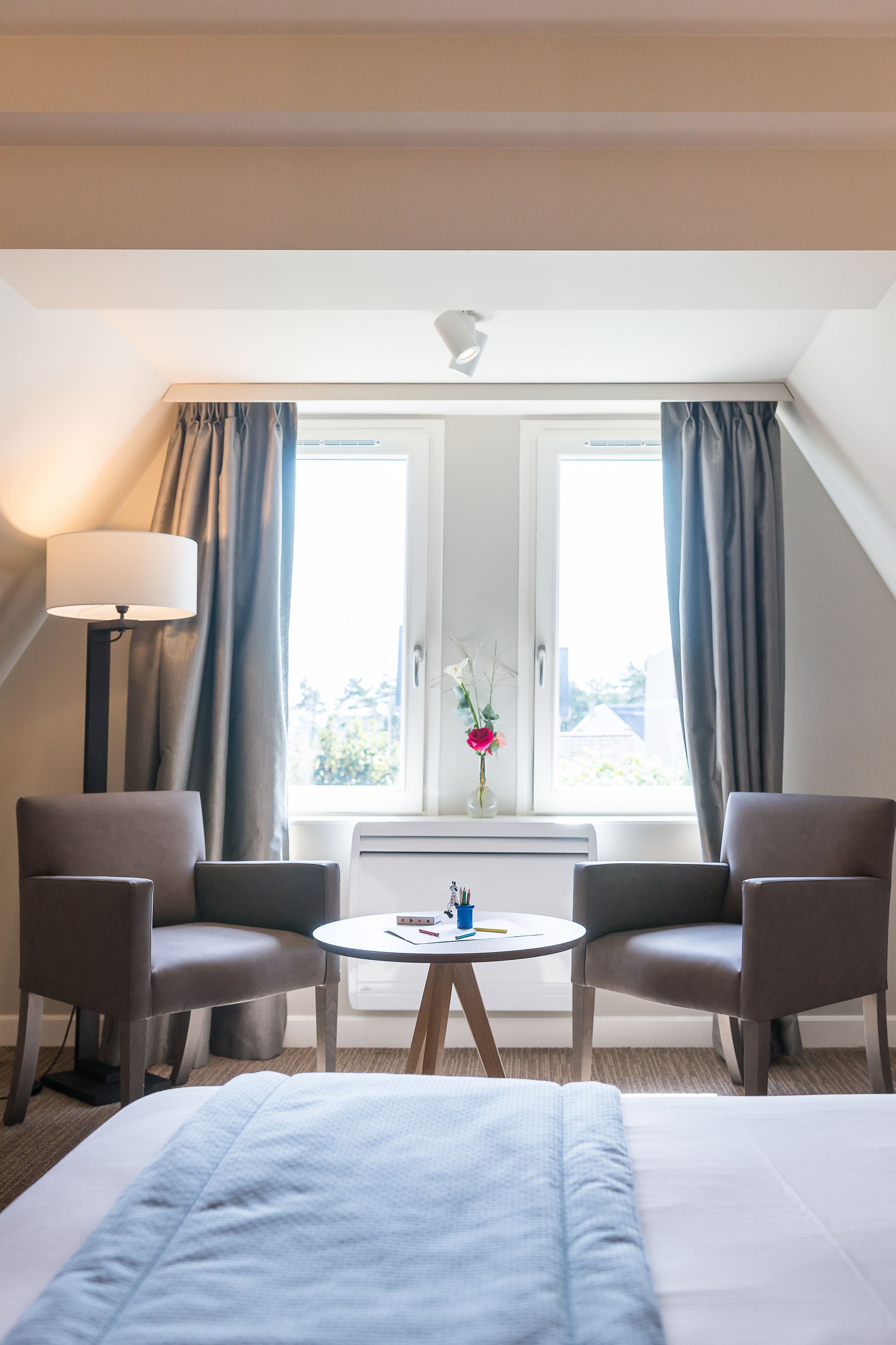 Une chambre d’hôtel avec un lit, deux fauteuils, une petite table, une lampe et une fenêtre avec des rideaux.