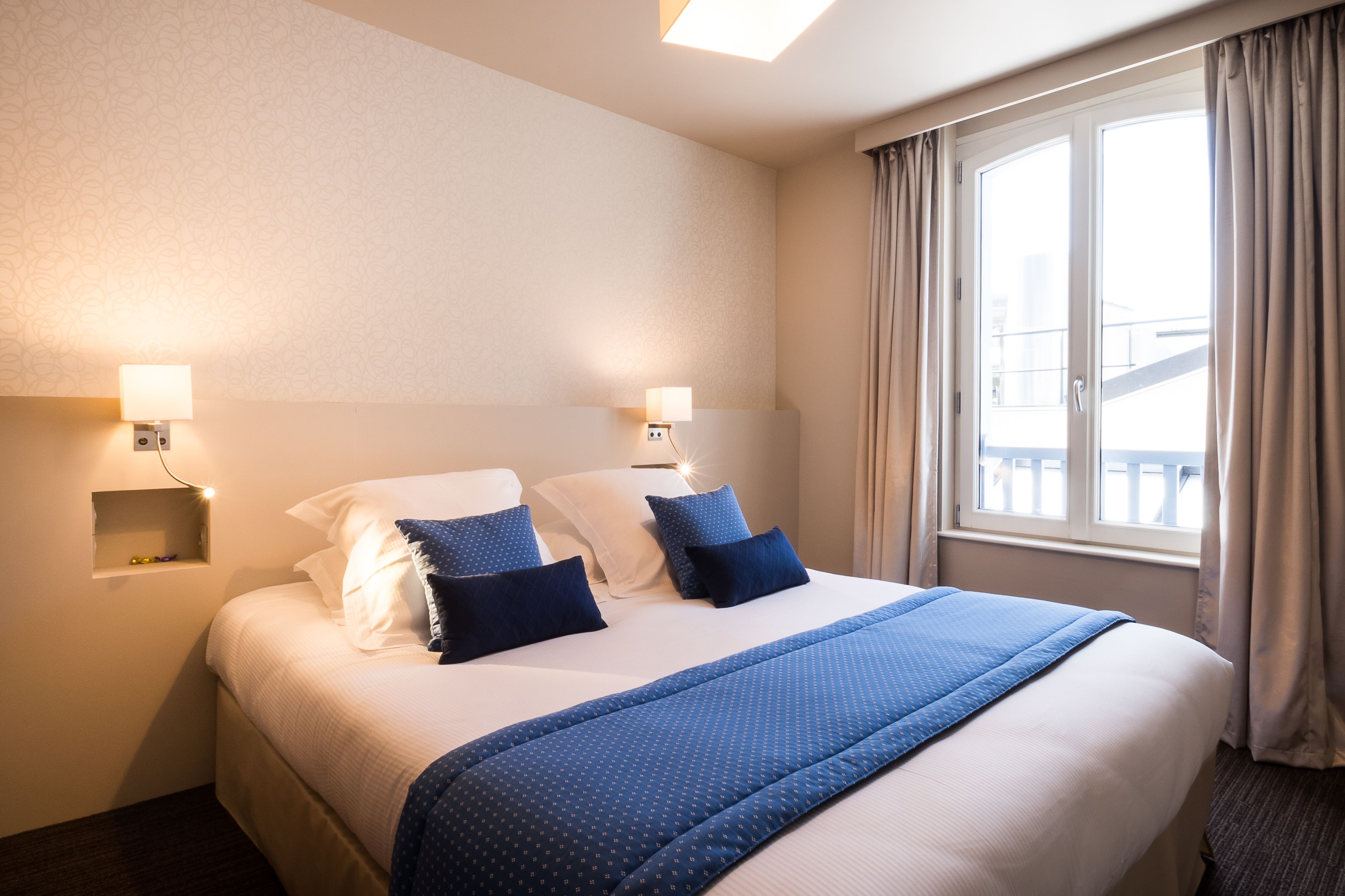 A hotel room with a large bed, two bedside lamps, a window with curtains, and a small shelf with decorative items.