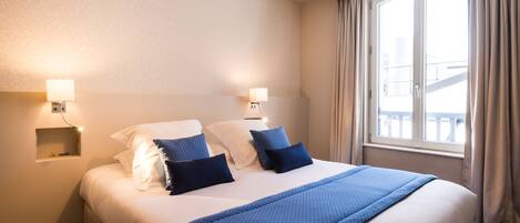 A hotel room with a large bed, two bedside lamps, a window with curtains, and a small shelf with decorative items.