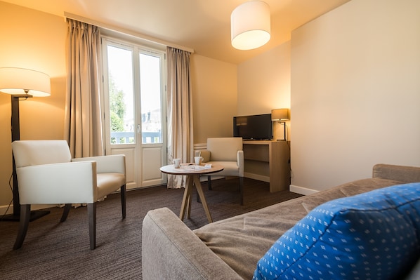Una habitación de hotel con un sofá, dos sillones, una mesita con una taza y un libro, un escritorio con un televisor, y una ventana con cortinas.