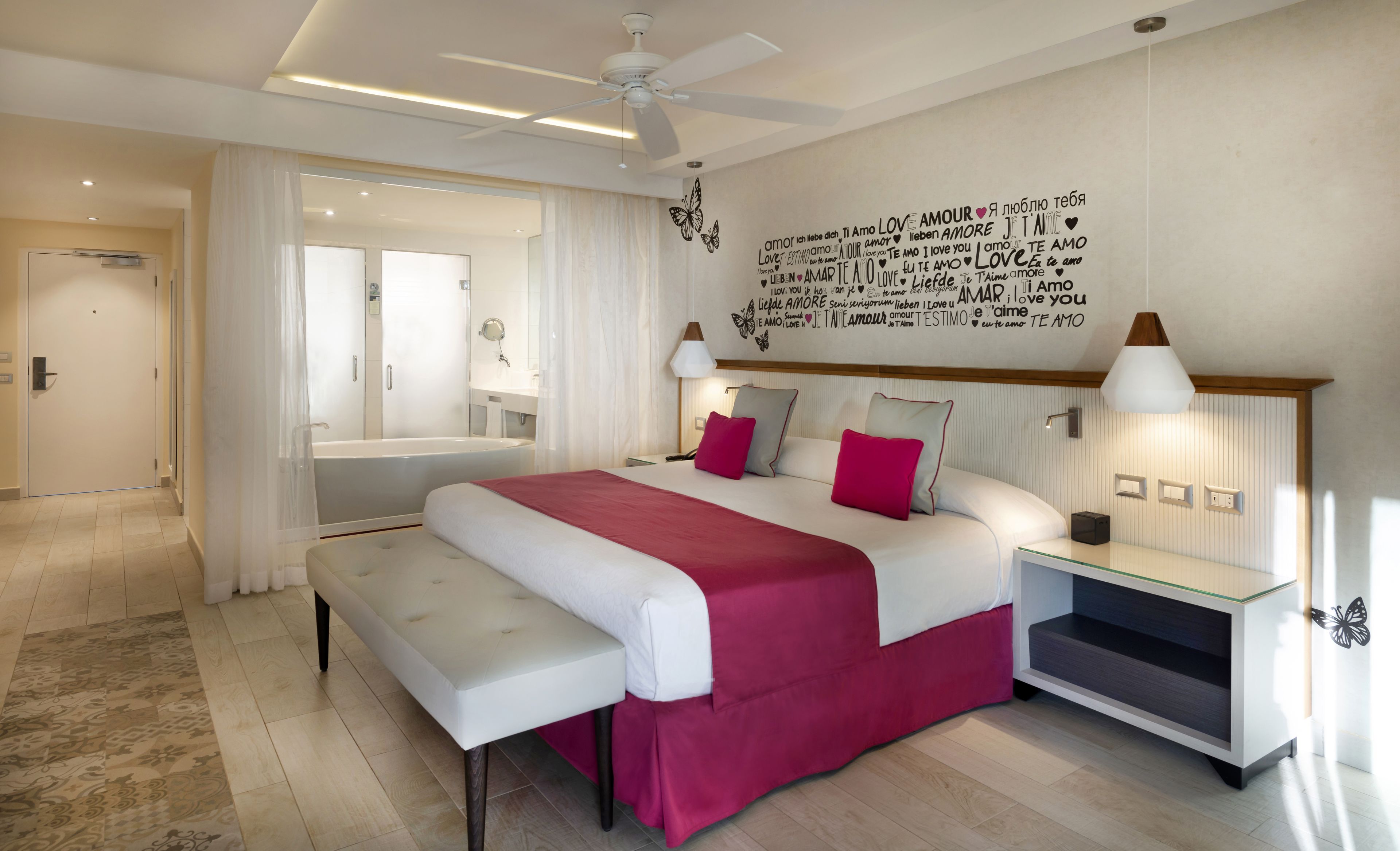 Une chambre d’hôtel moderne avec un grand lit, un banc, une table de chevet avec des lampes et un mur décoré sur le thème de l’amour.