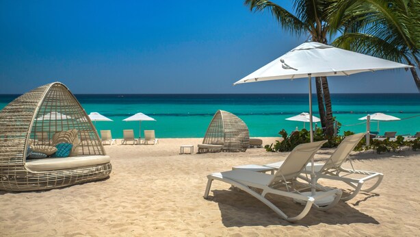 Una playa con sillas de playa blancas, una sombrilla blanca y una cabaña de mimbre.