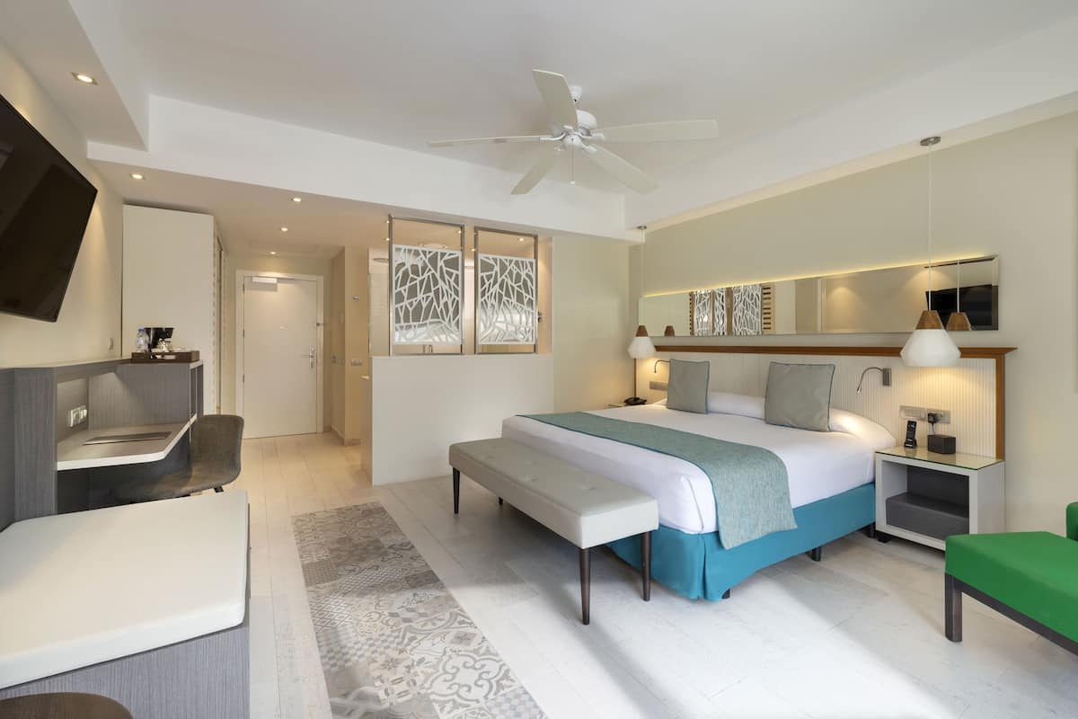 Ein modernes Hotelzimmer mit einem großen Bett, einem Schreibtisch, einem Sessel, einem Fernseher, einem Deckenventilator und einem Badezimmer mit Dusche und WC.