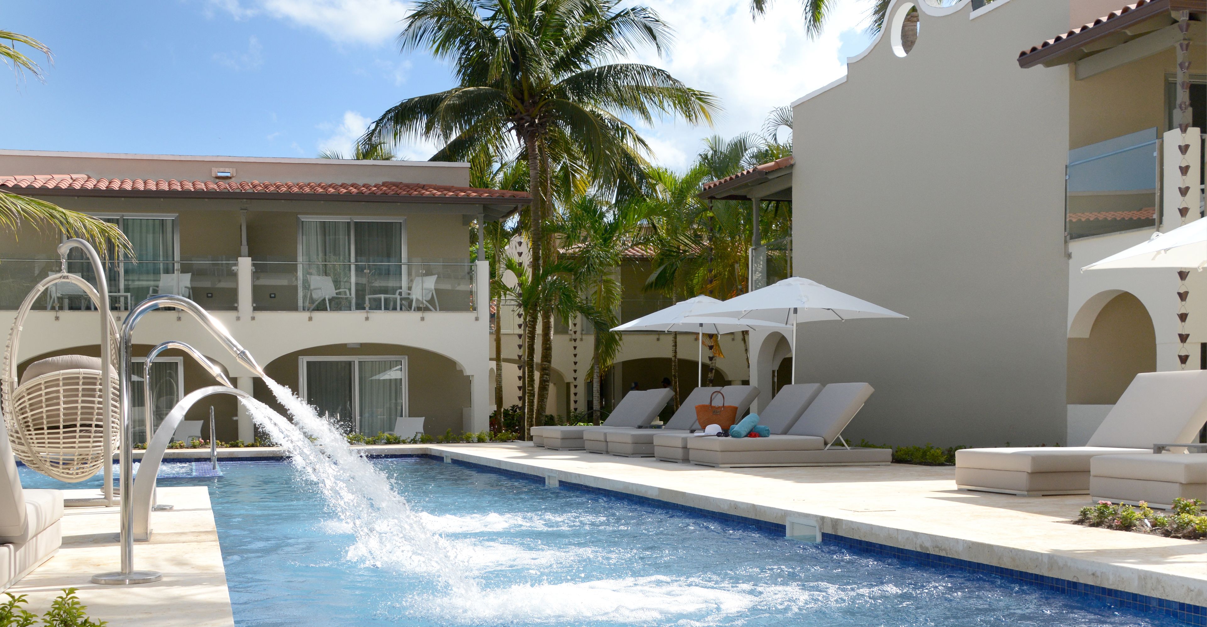 A pool area with lounge chairs and umbrellas, a water fountain, and a building with balconies and palm trees.
