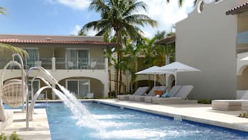 A pool area with lounge chairs and umbrellas, a water fountain, and a building with balconies and palm trees.