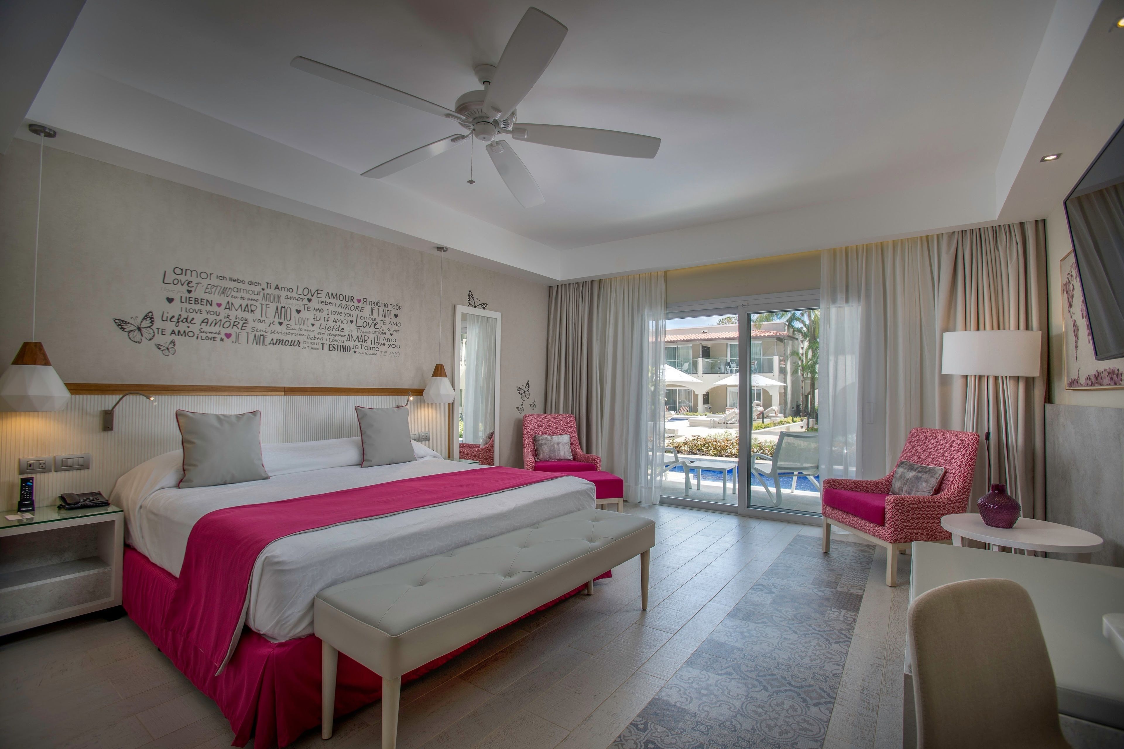 Habitación de hotel con una cama grande, ventilador de techo, escritorio con lámpara, una silla y vistas al exterior.