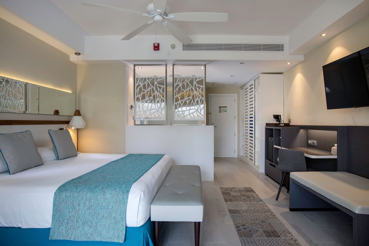Ein modernes Hotelzimmer mit Bett, Sitzbank, Schreibtisch, Fernseher und Deckenventilator.