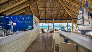Una terraza de madera con techo de paja que da al mar, con una zona de bar con asientos y un modelo de velero.