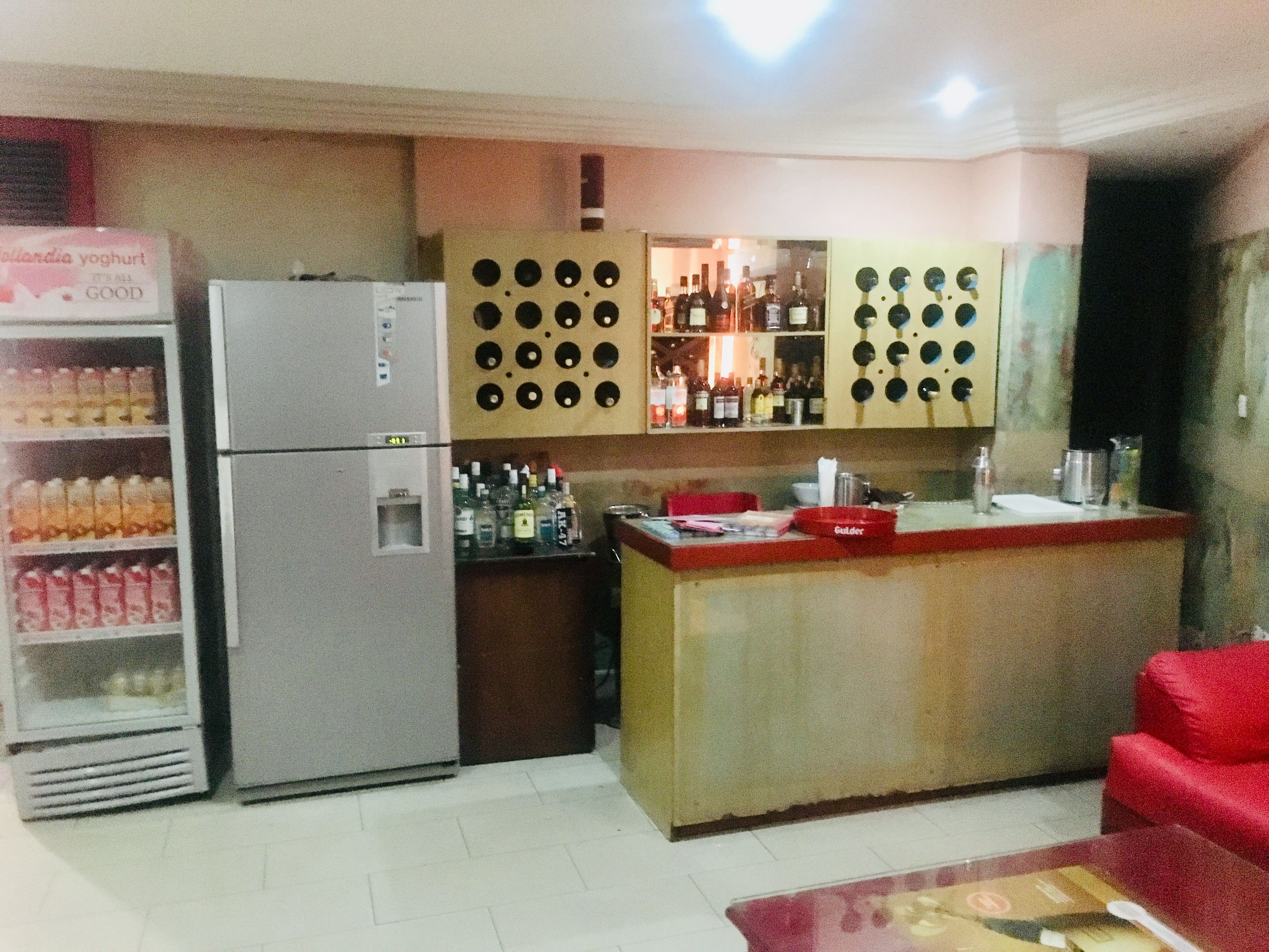 A compact kitchen with a refrigerator, a shelf of bottled drinks, a bar area with bottles, and a red couch.