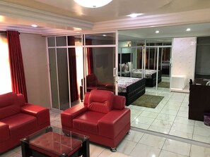 Un salon moderne avec des canapés en cuir rouge, une table basse en verre et une porte-fenêtre coulissante donnant sur une chambre à coucher équipée d’un lit et d’un bureau.