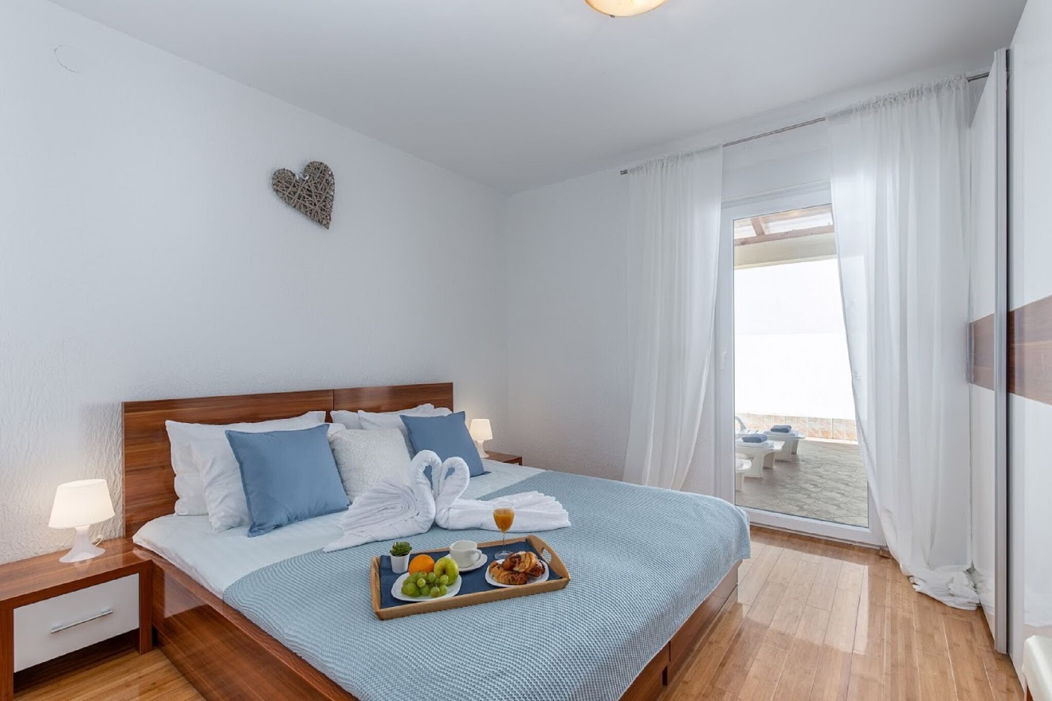 A neatly made bed with a tray of breakfast items, a heart-shaped decoration on the wall, and a view of the sea through the window.