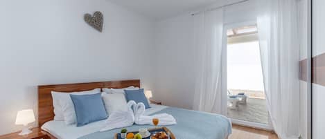 A neatly made bed with a tray of breakfast items, a heart-shaped decoration on the wall, and a view of the sea through the window.