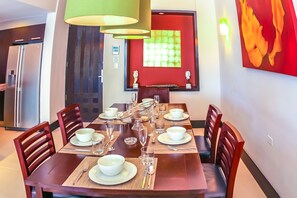 Un comedor con una mesa de madera dispuesta para seis personas, rodeada de sillas. Hay una cocina moderna con refrigerador y una mesa de comedor con una pared roja decorativa.