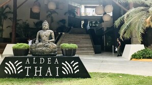 Una estatua de Buda frente a un edificio con el rótulo "ALDEA THAI", con una escalera que conduce a un nivel superior.