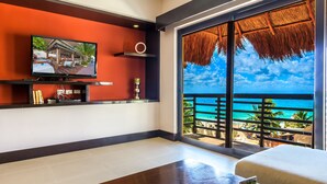 Una habitación con un ventanal grande que da a una playa, un televisor de pantalla plana en la pared y una mesita con un objeto decorativo.