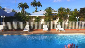 A pool area with white lounge chairs, a white fence, and palm trees in the background. - Domaine de May (Saint-François)