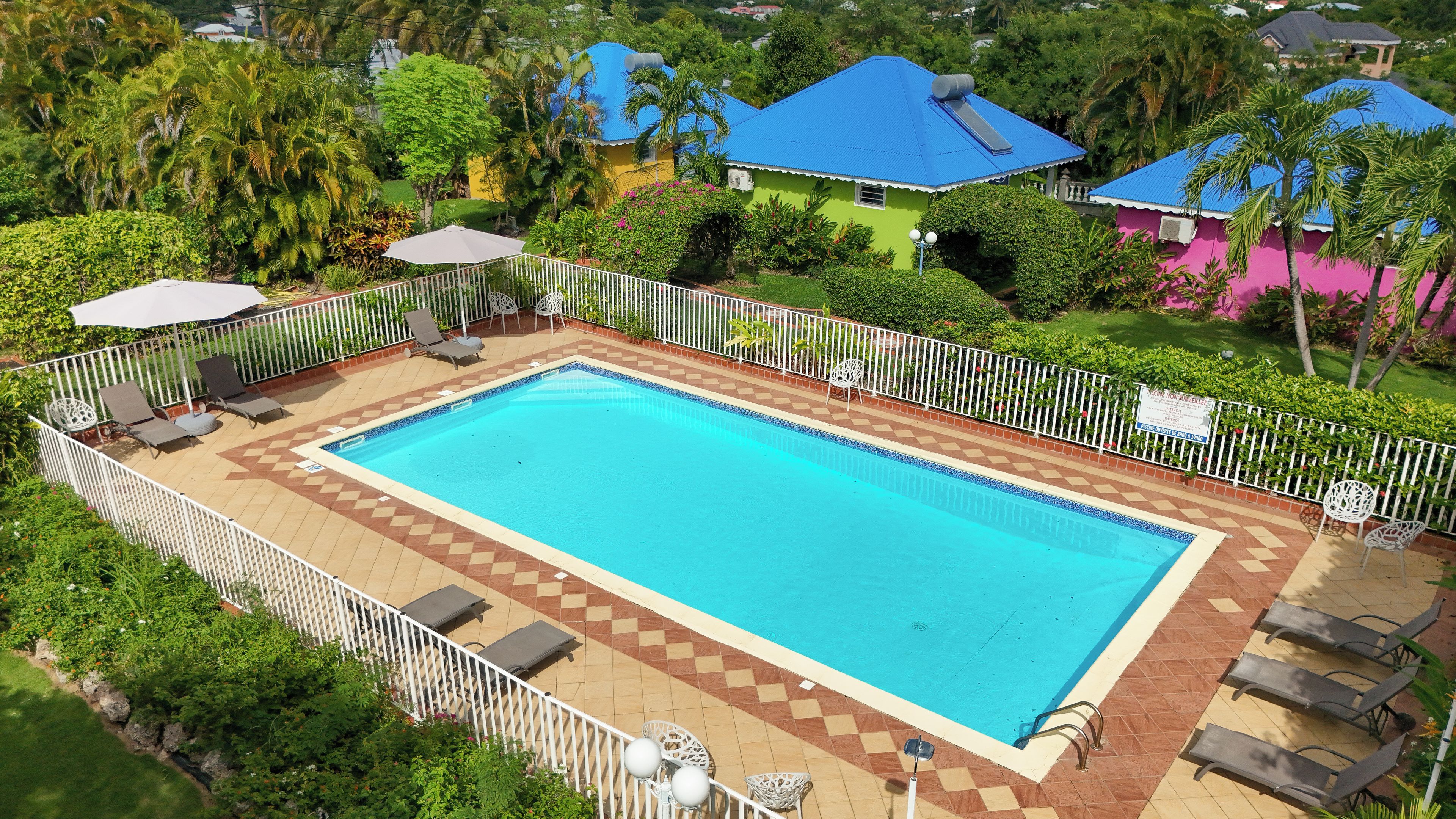A rectangular swimming pool with a tiled deck, surrounded by a white fence, and adjacent to a building with a blue roof.