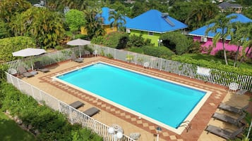 A rectangular swimming pool with a tiled deck, surrounded by a white fence, and adjacent to a building with a blue roof.